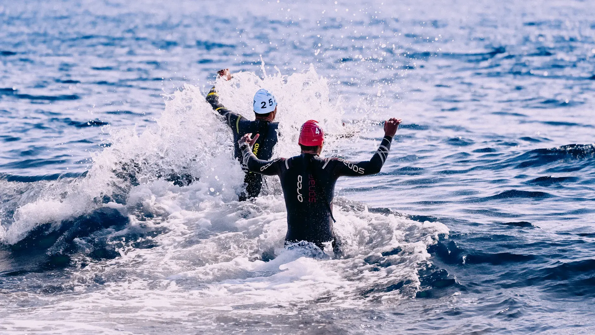 Dos nadadoras con gorro y traje de neoprene entrando al mar entre la espuma de una ola
